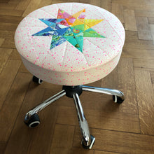 Load image into Gallery viewer, Rainbow Star Quilt Block Pattern by Penny Spool Quilts. Eight pointed star made using a rainbow of Tula Pink fabrics and used as a cover for wheeled office stool. Picture shows office stool from the side.