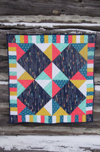 Load image into Gallery viewer, Canadian Diamond quilt pattern by Penny Spool Quilts, featuring four large diamonds surrounded by half-square triangles and a pieced border. Quilt shown in navy with pink, yellow, aqua and white accents, hanging from log wall.
