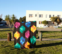 Load image into Gallery viewer, Facets quilt pattern by Penny Spool Quilts. Twin size quilt with large scale gemstones in pink, yellow, blue and green on black background, held up in front of house and palm trees.