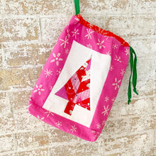 Load image into Gallery viewer, Festive Forest table runner and quilt block pattern by Penny Spool Quilts. Foundation paper pieced pattern. Image shows small drawstring bag made from a single block in pink, red and white, with a green drawstring.