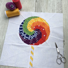 Load image into Gallery viewer, Swirly Lollipop FPP Block Pattern by Penny Spool Quilts. Foundation paper pieced quilt block pattern, sample is shown in rainbow colours with yellow and white striped stick, on white background. Shown with spools of thread and small scissors.
