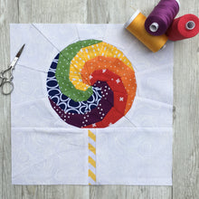 Load image into Gallery viewer, Swirly Lollipop FPP Block Pattern by Penny Spool Quilts. Foundation paper pieced quilt block pattern, sample is shown in rainbow colours wih yellow and white striped stick, on white background. Shown with spools of thread and small scissors.

