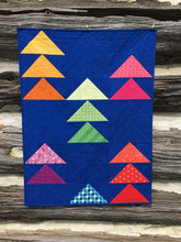 Load image into Gallery viewer, Flocks of Colour quilt pattern by Penny Spool Quilts. Baby quilt in rainbow flying geese on dark blue background, hanging on log wall.
