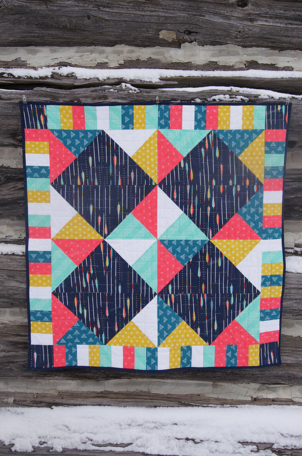 Canadian Diamond quilt pattern by Penny Spool Quilts, featuring four large diamonds surrounded by half-square triangles and a pieced border. Quilt shown in navy with pink, yellow, aqua and white accents, hanging from log wall.