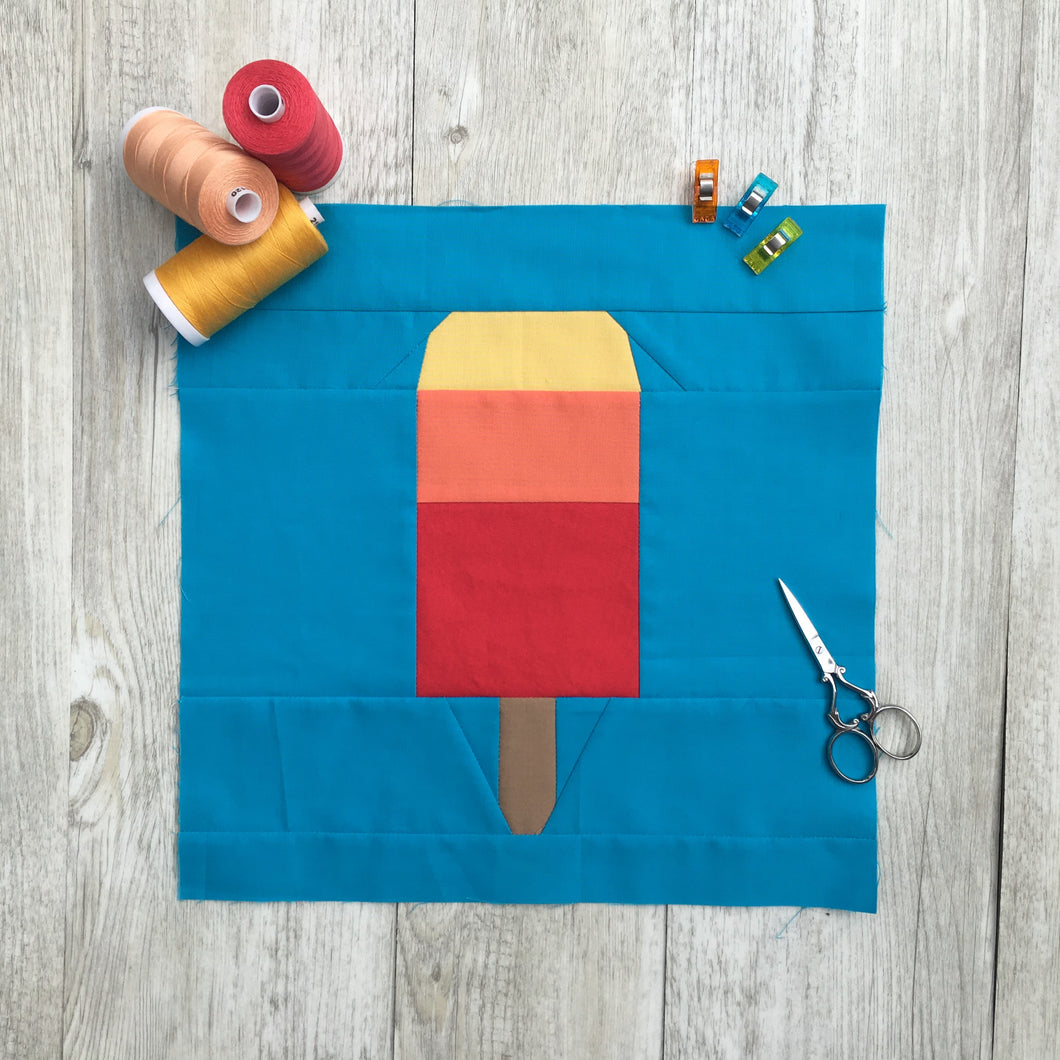 Popsicle 1 quilt block pattern by Penny Spool Quilts. Part of the Ice Cream Sunday collection. Tri-coloured popsicle in yellow, orange and red solid fabrics on blue background. shown with spools of thread, clips and small scissors
