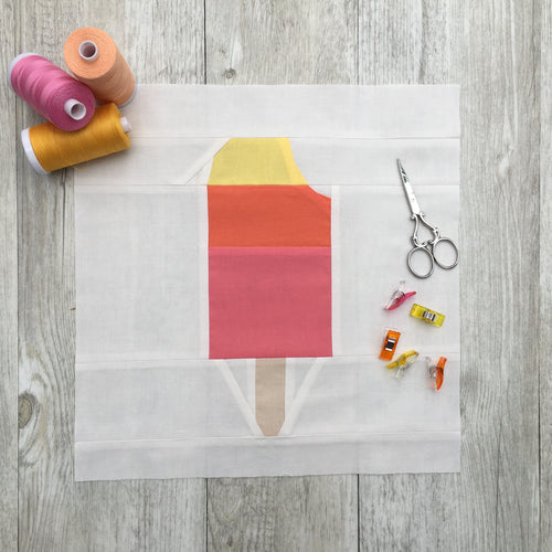 Popsicle 2 quilt block pattern by Penny Spool Quilts. Part of the Ice Cream Sunday collection. Tri-coloured popsicle with a bite taken out in top right corner, in yellow, orange and pink solid fabrics on white background. shown with spools of thread, clips and scissors
