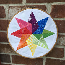 Load image into Gallery viewer, Rainbow Star Quilt Block Pattern by Penny Spool Quilts. Eight pointed star in rainbow colours on white background, shown in embroidery hoop in front of red brick wall
