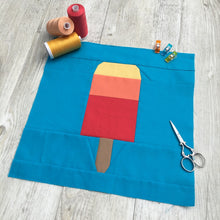 Load image into Gallery viewer, Popsicle 1 quilt block pattern by Penny Spool Quilts. Part of the Ice Cream Sunday collection. Tri-coloured popsicle in yellow, orange and red solid fabrics on blue background. shown with spools of thread, clips and scissors
